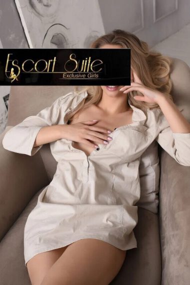 Woman reclining in a beige dress on a soft armchair in a modern room; a black 'Escort Suite Exclusive Girls' banner covers her eyes (advertisement).